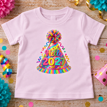 A ballerina pink short-sleeve baby t-shirt laid flat on a rustic wooden surface, decorated with colorful party props. The front of the shirt features a large graphic of a colorful party hat with 'Hello 2027' written on it in rainbow letters.