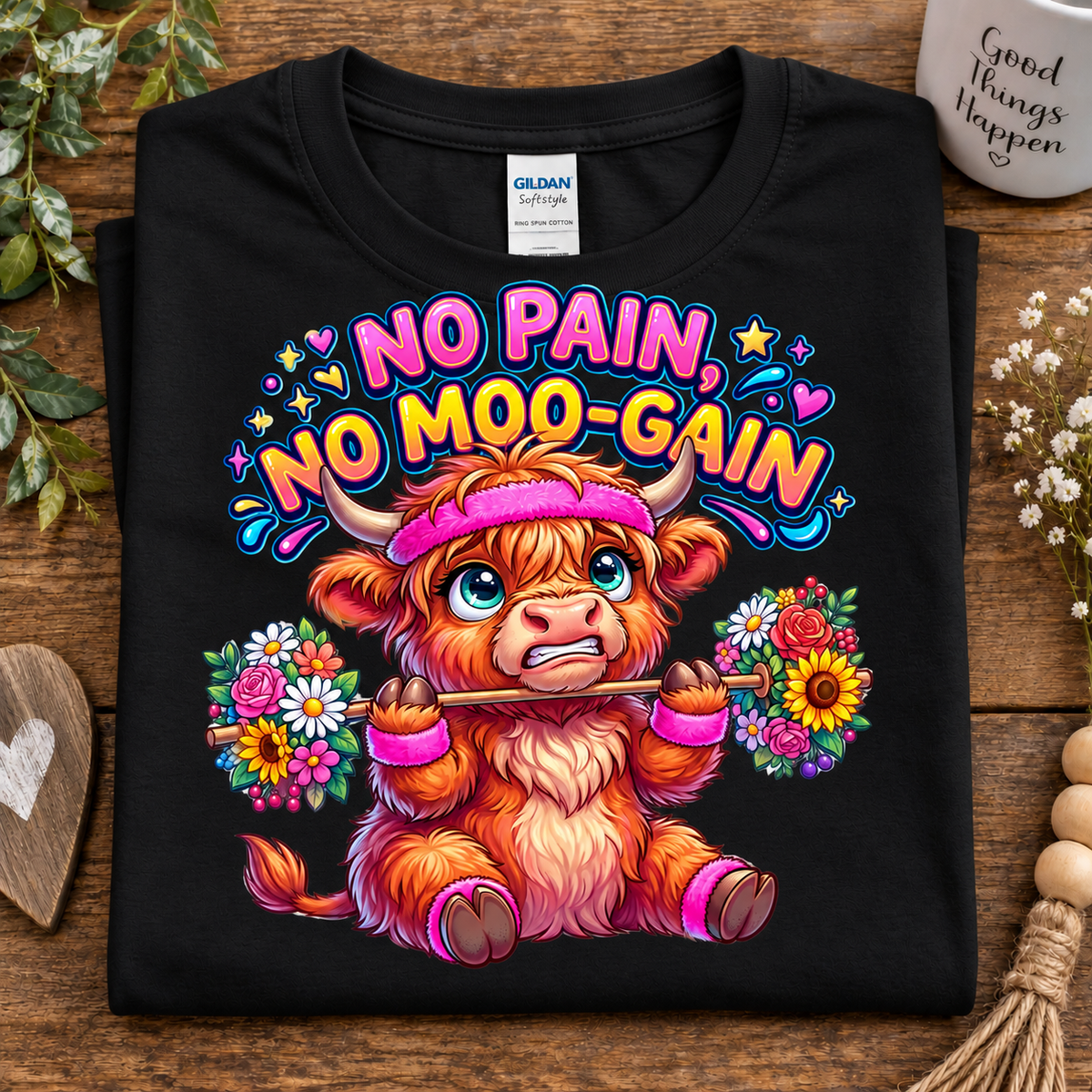 A folded black Gildan Comfort T-shirt is displayed on a rustic wooden surface. The shirt features a colorful graphic of a fluffy orange highland cow wearing a pink headband and holding flower bouquets, with the text 'NO PAIN NO MOO-GAIN' in vibrant pink, blue, and orange lettering.