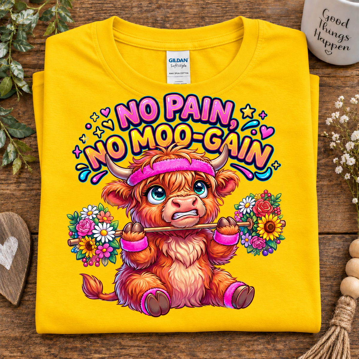 A folded daisy colored Gildan T-shirt on a rustic wooden surface, featuring a colorful cartoon highland cow graphic with the text 'NO PAIN, NO MOO-GAIN' and floral accents.