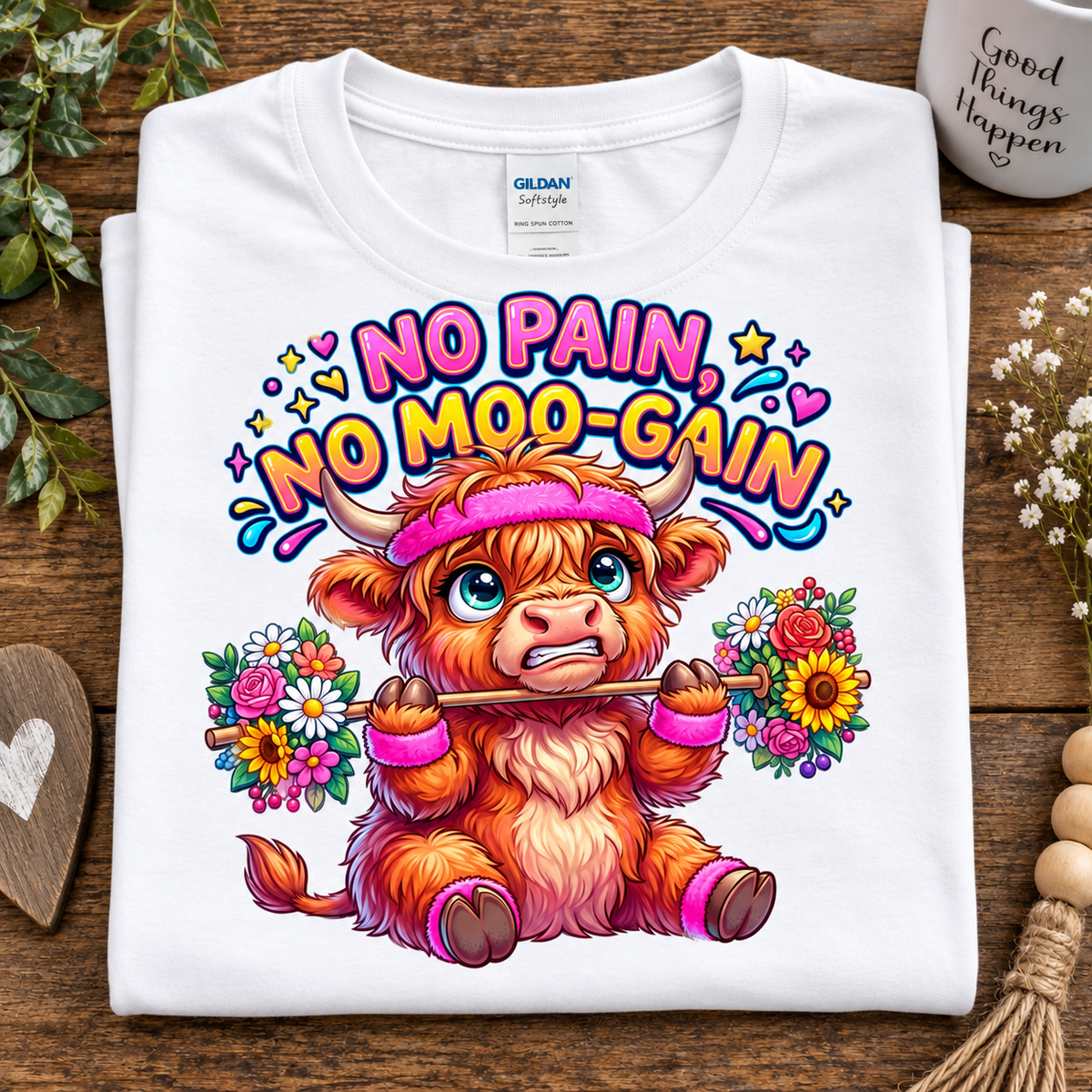 A folded white Gildan Softstyle T-shirt with a colorful graphic of a fluffy orange-and-white cartoon cow wearing a pink headband and holding flowers, under the text 'NO PAIN, NO MOO-GAIN.' The shirt is styled on a wooden surface with decorative flowers, a heart-shaped coaster, and a mug in the background.
