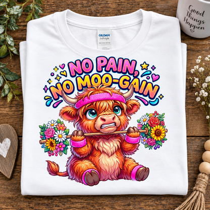 A folded white Gildan Softstyle T-shirt with a colorful graphic of a fluffy orange-and-white cartoon cow wearing a pink headband and holding flowers, under the text 'NO PAIN, NO MOO-GAIN.' The shirt is styled on a wooden surface with decorative flowers, a heart-shaped coaster, and a mug in the background.