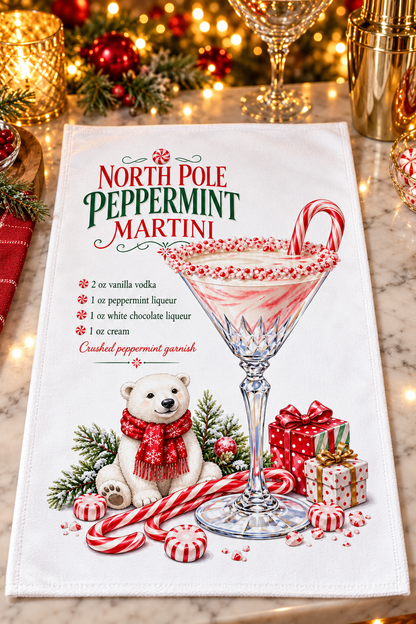 A white kitchen towel featuring a festive Christmas design for a 'North Pole Peppermint Martini' with a cocktail illustration, a polar bear, candy canes, and gift boxes, laid flat on a marble surface with warm holiday lights in the background.