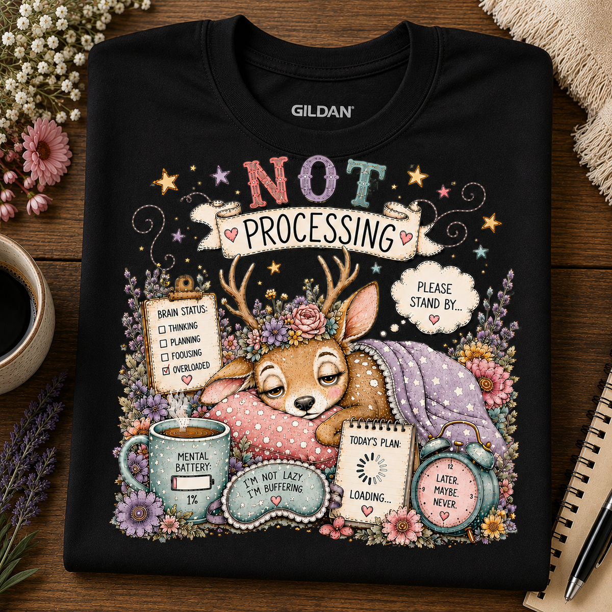 A black Gildan crewneck t-shirt with a colorful graphic print of a sleeping deer surrounded by flowers, a coffee mug, and text that reads 'NOT PROCESSING'. The shirt is displayed flat on a wooden table with decorative props like a coffee cup, flowers, and a notebook.