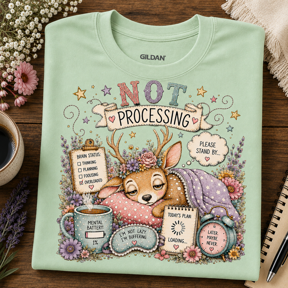 A mint green Gildan t-shirt with a detailed cartoon graphic of Bambi sleeping under a floral crown, surrounded by humorous text bubbles like 'NOT PROCESSING' and 'BRAIN ENERGY 10%'. The shirt is laid flat on a dark wooden table surrounded by decorative props including flowers, a coffee cup, and a notebook.