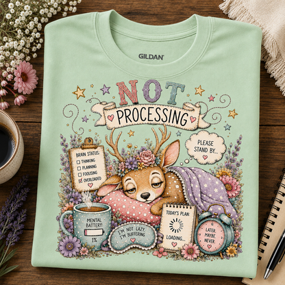 A mint green Gildan t-shirt with a detailed cartoon graphic of Bambi sleeping under a floral crown, surrounded by humorous text bubbles like 'NOT PROCESSING' and 'BRAIN ENERGY 10%'. The shirt is laid flat on a dark wooden table surrounded by decorative props including flowers, a coffee cup, and a notebook.