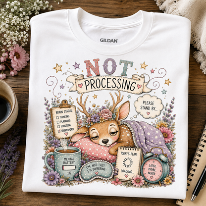 A white Gildan crewneck t-shirt laid flat on a rustic wooden table, featuring a detailed graphic of a sleeping deer surrounded by flowers and humorous text like 'NOT PROCESSING' and 'PLEASE STAND BY.'.