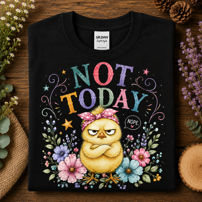 A black Gildan Sof-tstyle t-shirt laid flat on a rustic wooden surface, surrounded by decorative flowers, greenery, and a wood slice. The shirt features a colorful graphic of a grumpy yellow chick wearing a pink bow headband, with the text 'NOT TODAY' and 'NOPE' surrounded by floral illustrations.