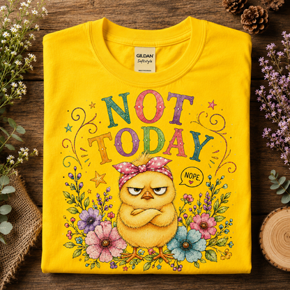 A folded daisy colored Gildan Softstyle t-shirt with a colorful graphic of an angry yellow chick wearing a pink polka-dot bow headband, surrounded by flowers and the text 'NOT TODAY' and 'NOPE', displayed on a rustic wooden background with decorative greenery.
