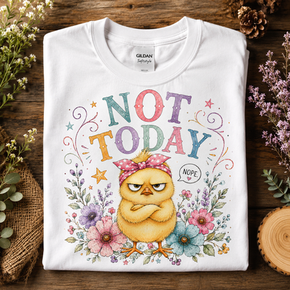A folded white Gildan Softstyle t-shirt with a colorful graphic of a grumpy yellow chick under the text 'NOT TODAY' surrounded by flowers, displayed on a rustic wooden background with decorative plants.