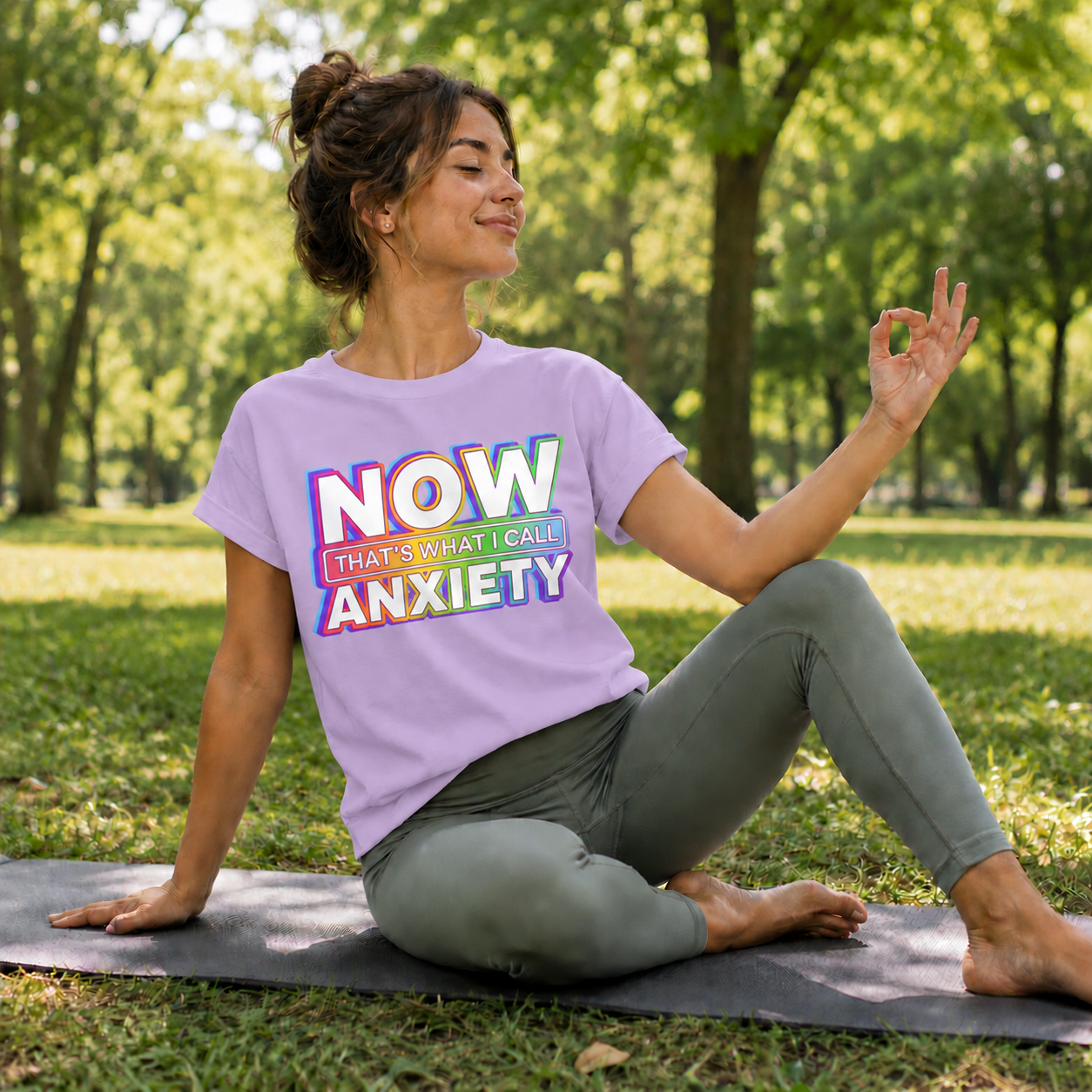 Woman sitting on a yoga mat in a park wearing a t-shirt with colorful text.