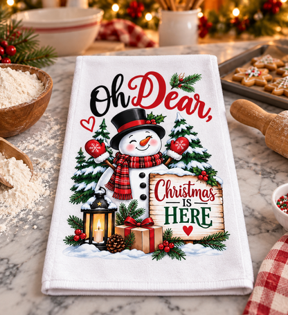 A white kitchen tea towel with a festive Christmas print, featuring a snowman, pine trees, a lantern, and the text 'Oh Dear, Christmas is Here'. The towel is displayed on a marble countertop surrounded by holiday baking props like flour, a rolling pin, and a tray of cookies.