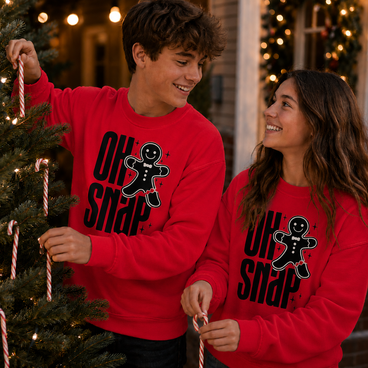 A man and a woman wearing matching red crewneck sweatshirts with a black gingerbread man graphic and the text 'OH SNAP' hang candy canes next to a decorated Christmas tree in a festive home setting.