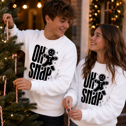 A man and a woman wearing matching white crewneck sweatshirts with a black 'OH SNAP' gingerbread man graphic, posing with candy canes next to a decorated Christmas tree in a festive indoor setting.