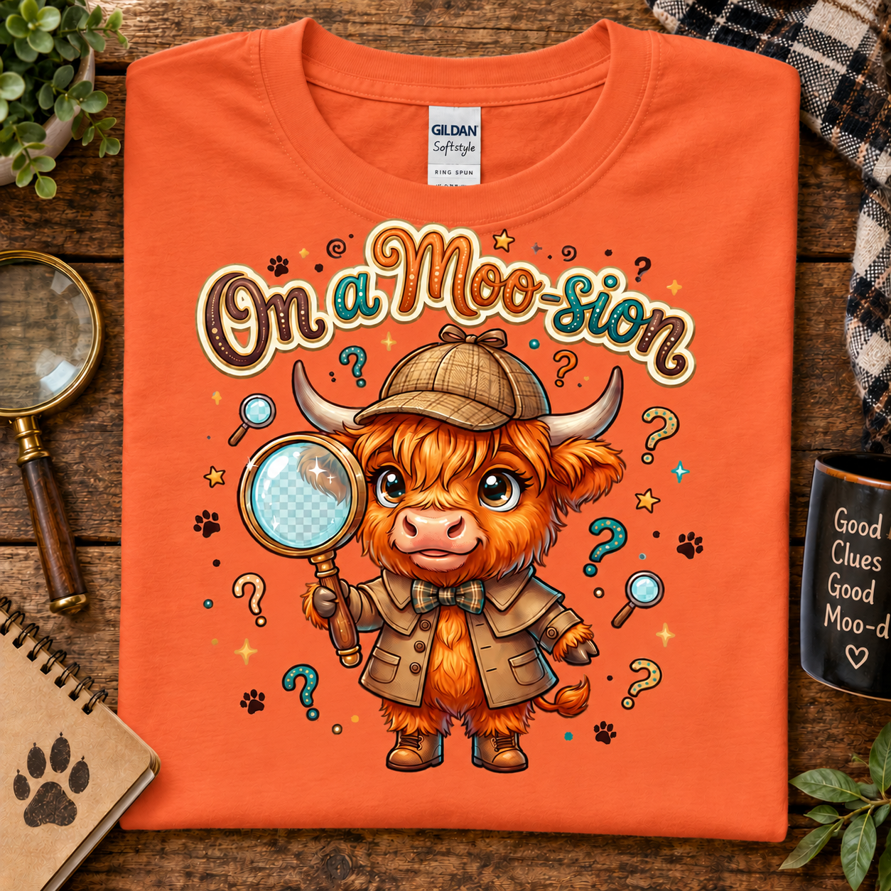 An orange t-shirt laid flat on a rustic wooden table, featuring a cartoon illustration of a highland cow dressed as a detective with the text 'On a Moo-sion'. The shirt is styled with a magnifying glass, notebook, and a mug in the background.