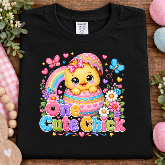 A black short-sleeve t-shirt laid flat on a rustic wooden surface, decorated with Easter-themed props like pink eggs, flowers, and beads. The shirt features a colorful graphic of a cute cartoon chick in a striped Easter egg with a rainbow and the text 'One Cute Chick' in pastel rainbow letters.
