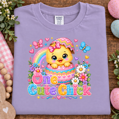 A lavender purple t-shirt with a colorful Easter graphic of a cute cartoon chick in a basket, surrounded by flowers, butterflies, and the text 'One Cute Chick'. The t-shirt is displayed in a flat-lay arrangement on a wooden surface with decorative Easter eggs, flowers, and a gingham cloth.