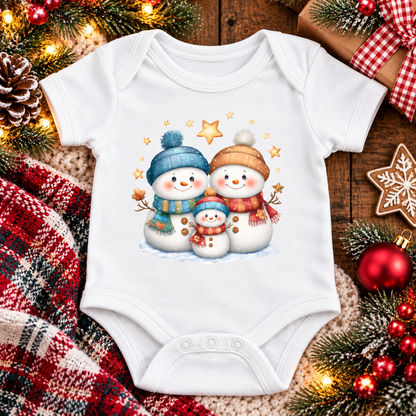 A white short-sleeve baby bodysuit laid flat on a rustic wooden surface, decorated with a colorful illustration of three snowmen wearing winter hats and scarves. The bodysuit is surrounded by holiday-themed props including a red plaid blanket, pine branches with fairy lights, red ornaments, and wrapped gift boxes.