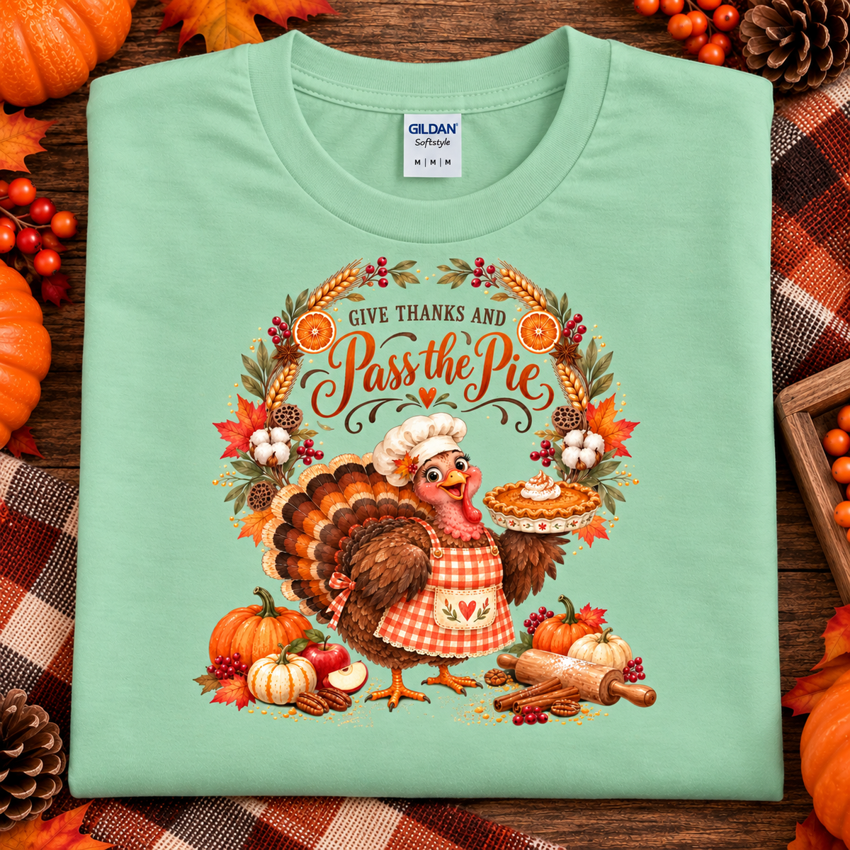 A folded mint green Gildan Softstyle t-shirt with a detailed Thanksgiving graphic featuring a turkey chef and a pie, surrounded by autumn elements like pumpkins, leaves, and a pie, displayed on a rustic wooden background with plaid fabric and seasonal decorations.