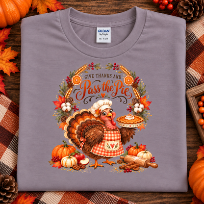 A folded Gildan Softstyle t-shirt in paragon purple, featuring a colorful Thanksgiving graphic of a cartoon turkey holding a pie next to a pie crust, with the text 'GIVE THANKS AND Pass the Pie'. The shirt is styled on a rustic wooden background with autumn leaves, pumpkins, and a plaid blanket.