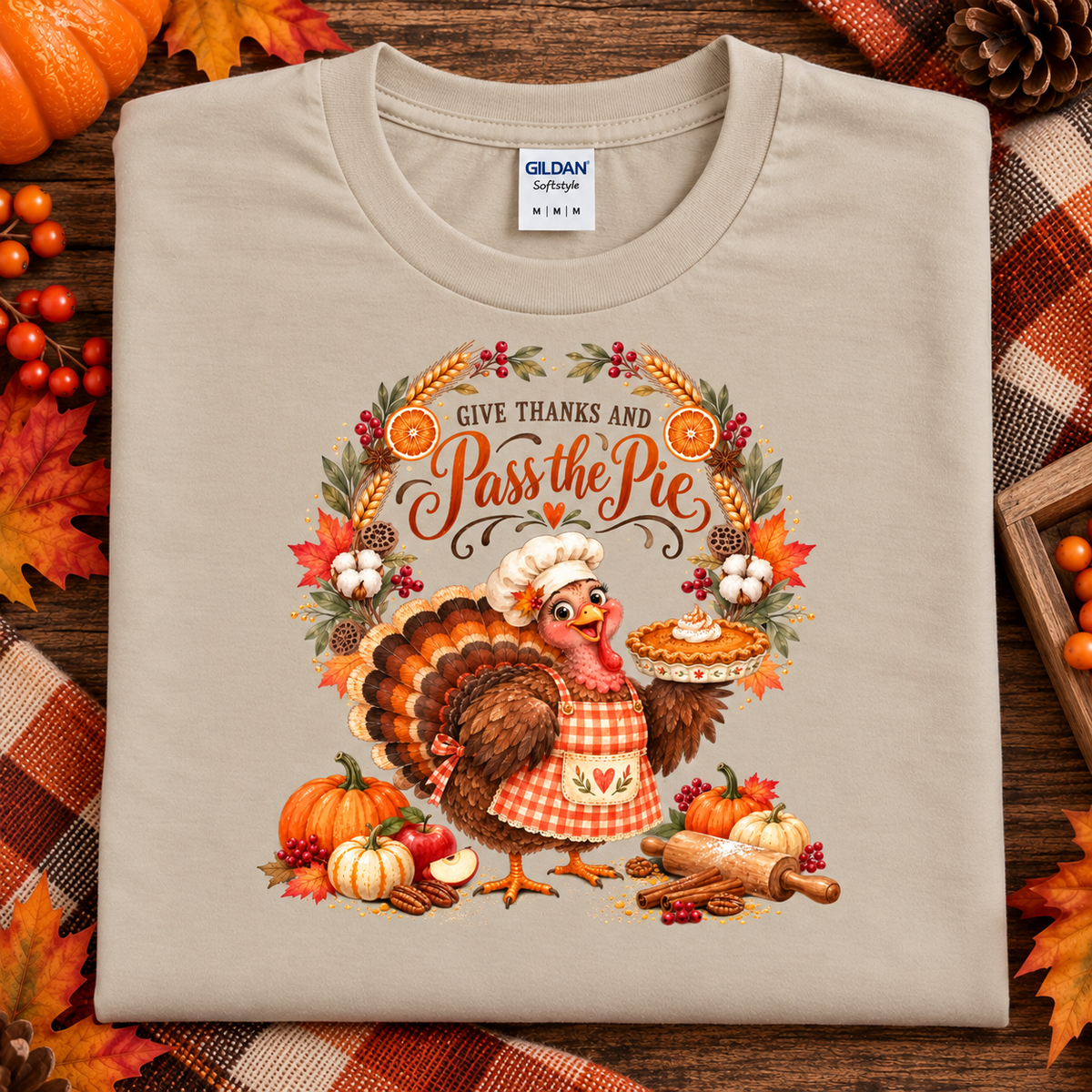 A folded sand colored Gildan Softstyle t-shirt with a detailed Thanksgiving graphic featuring a turkey chef and a pie, surrounded by autumn elements like pumpkins, leaves, and a pie, displayed on a rustic wooden background with plaid fabric and seasonal decorations.