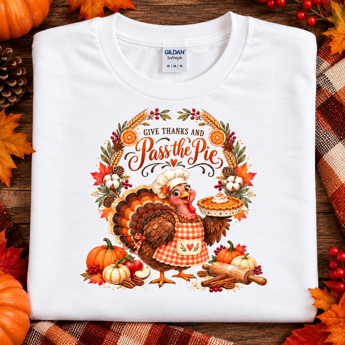 A folded white Gildan Comfortstyle T-shirt with a colorful Thanksgiving graphic reading 'Give Thanks and Pass the Pie,' featuring a turkey, pumpkins, autumn leaves, and pie. The shirt is styled on a wooden surface with fall leaves, pumpkins, and plaid fabric around it.