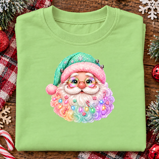 A folded keylime green crewneck t-shirt featuring a colorful graphic of a cute, smiling Santa Claus with a fluffy multicolored beard and a pink and green hat, displayed on a rustic wooden background with Christmas decorations.