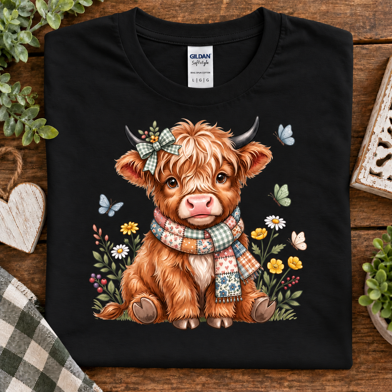 A folded black Gildan Softstyle t-shirt with a cute cartoon Highland cow graphic, styled on a rustic wooden background with decorative plants and a heart-shaped ornament.