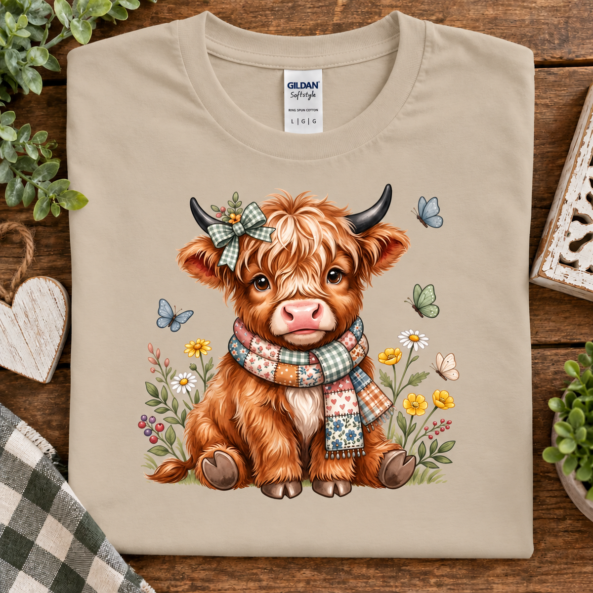 A folded sand colored Gildan Softstyle t-shirt with a colorful graphic of a fluffy brown highland cow wearing a plaid bow and scarf, surrounded by flowers, butterflies, and greenery, displayed on a rustic wooden background.