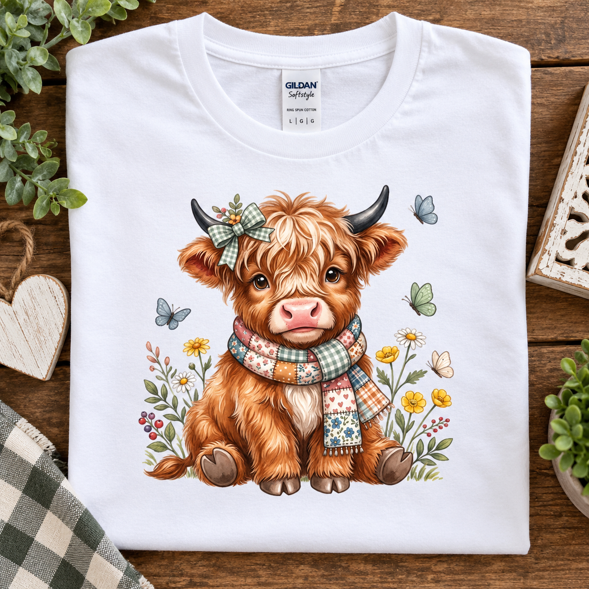 A folded white Gildan Softstyle T-shirt with a colorful cartoon illustration of a fluffy brown Highland cow wearing a plaid bow and scarf, surrounded by flowers and butterflies, displayed on a wooden surface with decorative props.