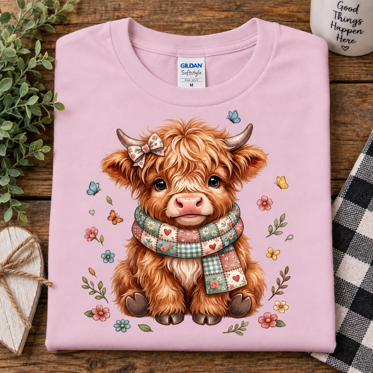 A folded light pink Gildan Softstyle T-shirt with a cute cartoon Highland cow graphic, displayed on a rustic wooden surface with decorative props.