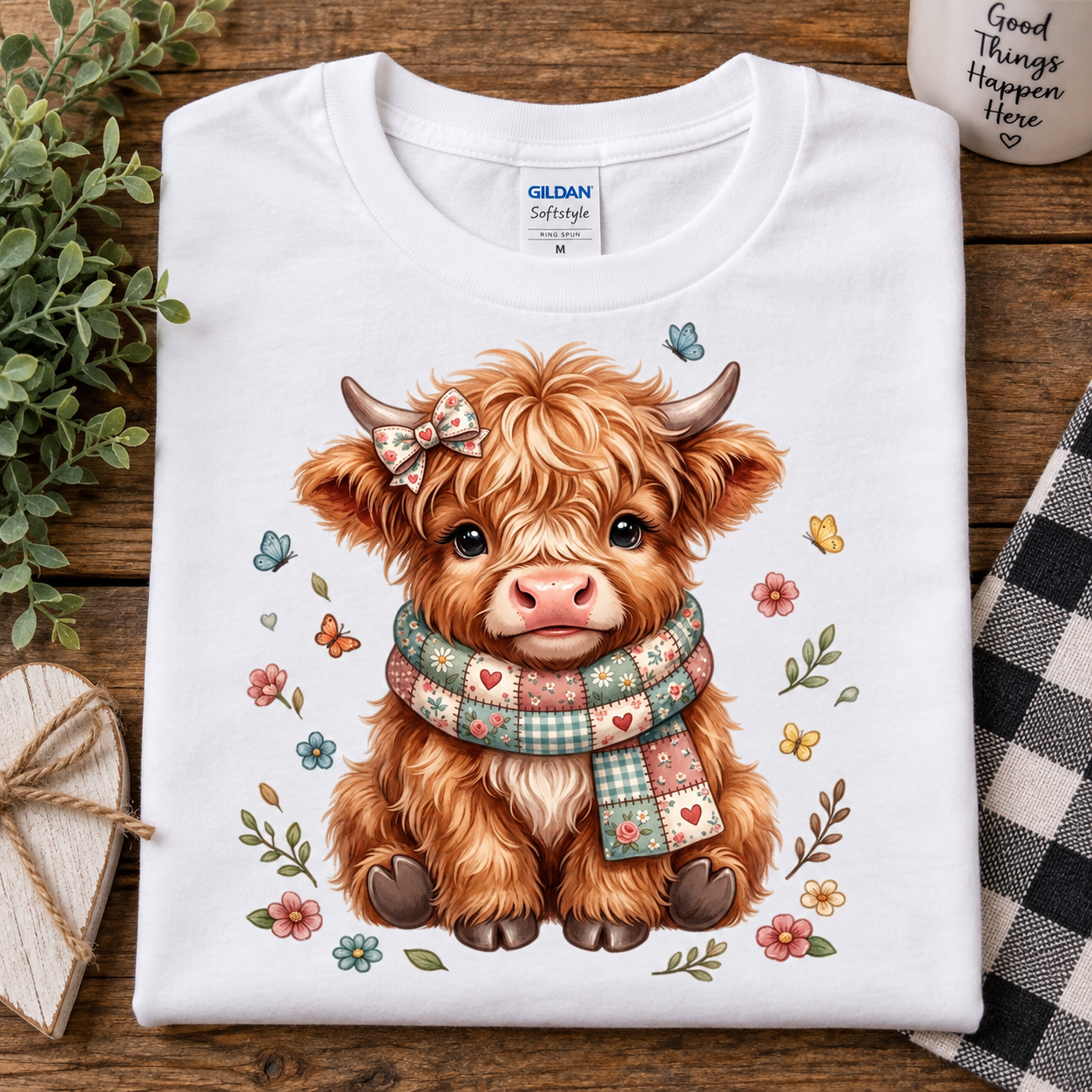 A folded white Gildan Softstyle T-shirt with a cute cartoon Highland cow illustration, shown on a rustic wooden surface with decorative plants, a mug, and a checkered cloth.