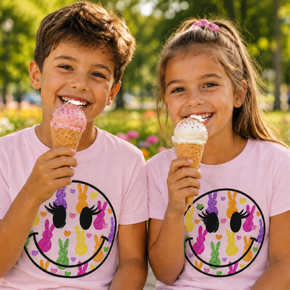 Two children enjoying ice cream cones in a park with a smiley face graphic design on their light pink shirts.