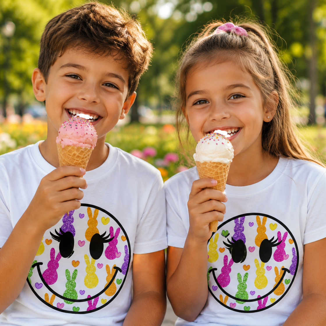 Two children enjoying ice cream cones in a park with a smiley face graphic design on their white shirts.