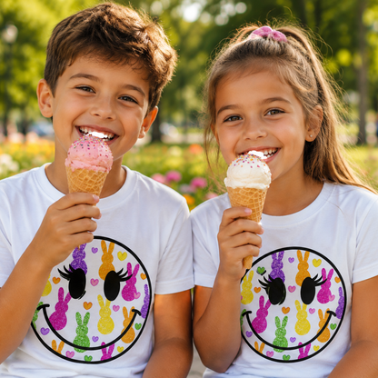 Two children enjoying ice cream cones in a park with a smiley face graphic design on their white shirts.