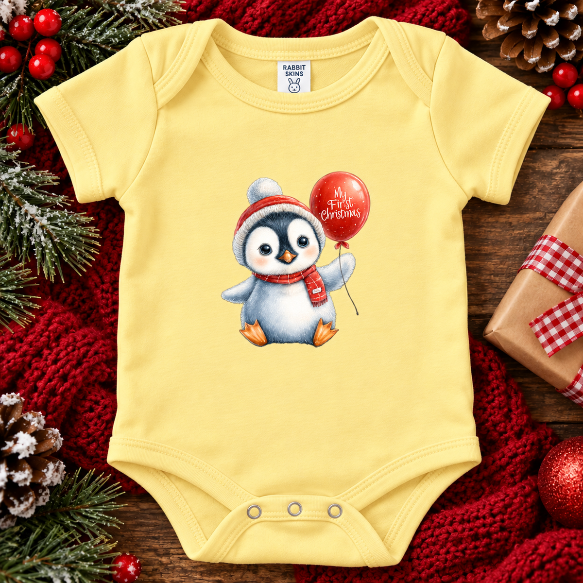 A butter yellow short-sleeve baby bodysuit laid flat on a rustic wooden surface, decorated with a penguin illustration holding a red Christmas balloon. The bodysuit is surrounded by festive holiday props including a red knit blanket, pine branches, red berries, and a wrapped gift box.