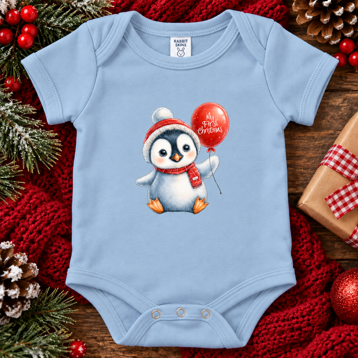 A light blue baby bodysuit with a penguin graphic holding a red balloon that says 'My First Christmas'. The bodysuit is styled in a flat-lay on a wooden surface with a red knit blanket, pine branches, pinecones, and a wrapped gift box.
