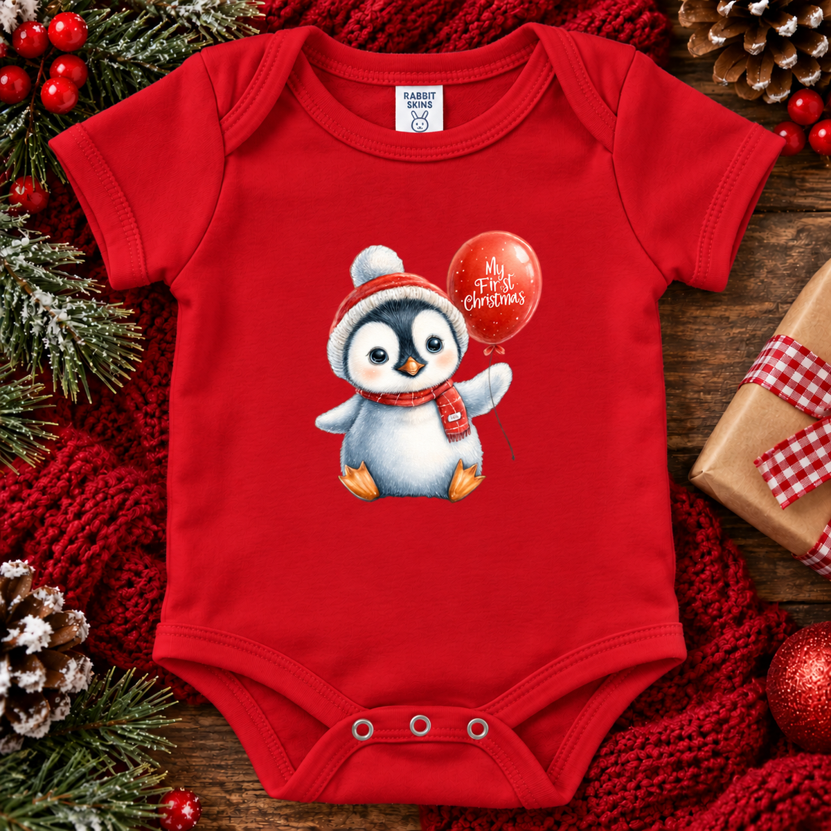 A red baby bodysuit with a cartoon penguin wearing a Santa hat and scarf while holding a red balloon that says 'My First Christmas.' The bodysuit is laid flat on a rustic wooden surface surrounded by holiday decorations including pine branches, red berries, a red knit blanket, and a wrapped gift box.