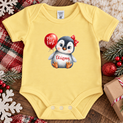 A butter yellow baby bodysuit laid flat on a wooden surface with holiday decorations around it. The bodysuit has a printed graphic of a penguin holding a red balloon that says 'My first Christmas' with 'Christmas' written in red script on the belly.