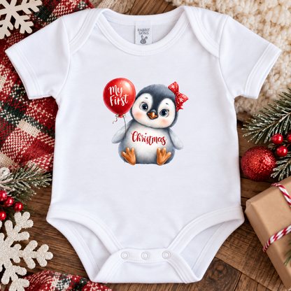 A white baby bodysuit laid flat on a rustic wooden surface, decorated with a cute penguin graphic holding a red balloon that says 'my first Christmas.' The bodysuit is surrounded by holiday props including a plaid blanket, pine branches, red ornaments, and a wrapped gift box.
