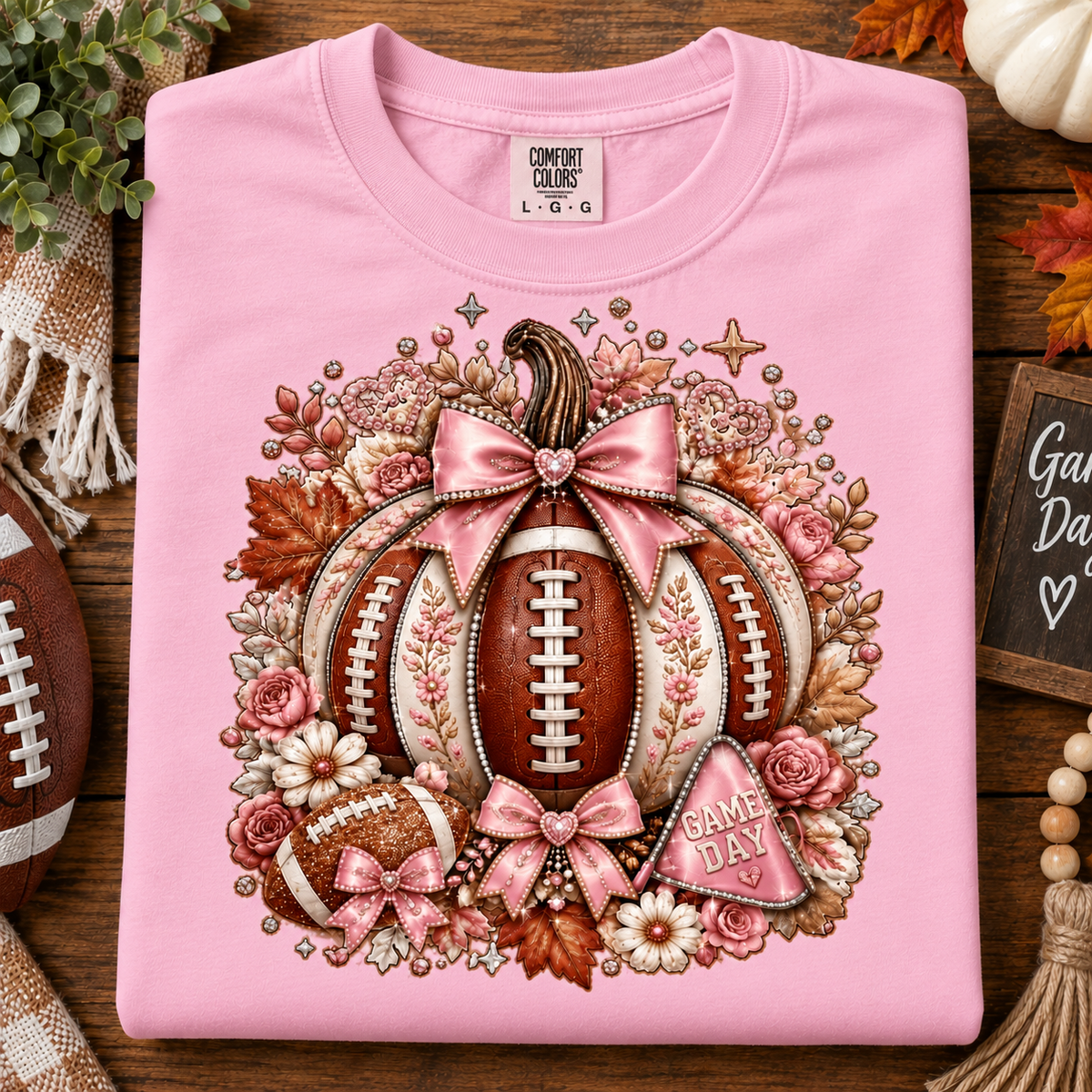 A folded light pink t-shirt with a detailed autumn football graphic, displayed on a rustic wooden surface with seasonal props like a partial football, a pumpkin, and fall leaves.