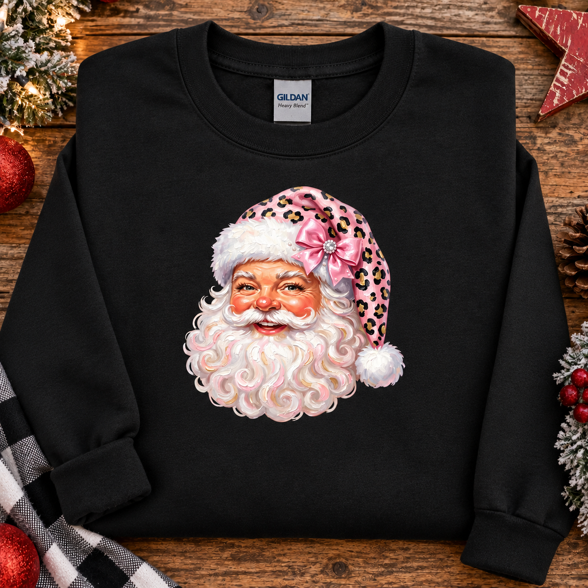 A black crewneck sweatshirt featuring a graphic of a smiling Santa Claus face with a pink leopard-print hat and bow, laid flat on a rustic wooden surface with holiday decorations.