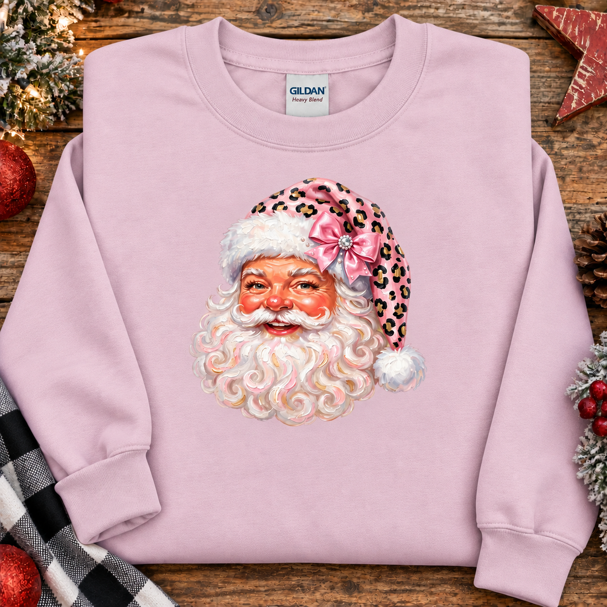 A light pink crewneck sweatshirt laid flat on a rustic wooden surface, featuring a large graphic of Santa Claus's face wearing a pink leopard-print Santa hat with a bow. The sweatshirt is styled with holiday decorations including a red ornament, pinecones, a plaid blanket, and a star ornament.