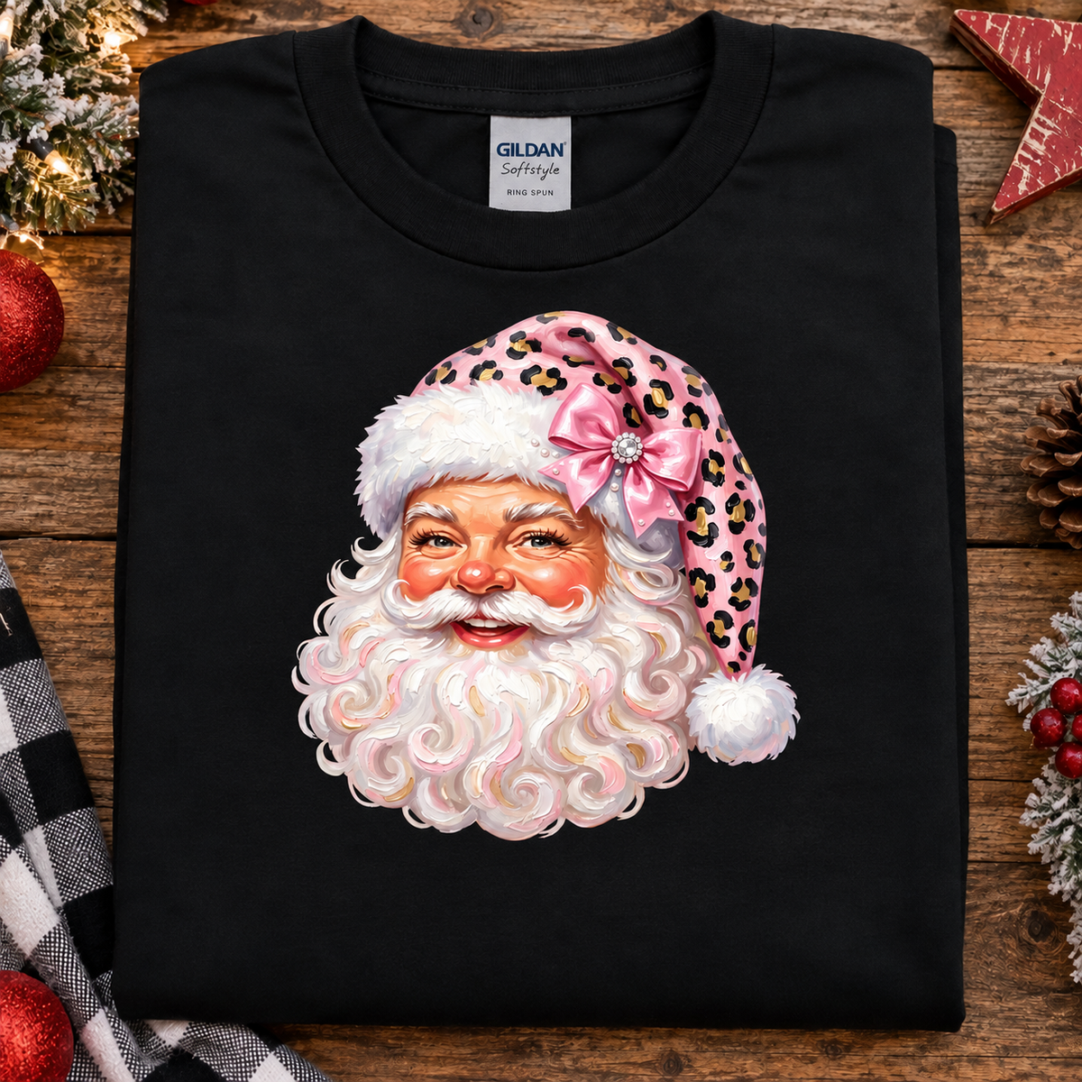 A folded black Gildan Softstyle t-shirt with a graphic of a Santa Claus face wearing a pink leopard-print hat with a bow, displayed on a rustic wooden background with holiday decorations.