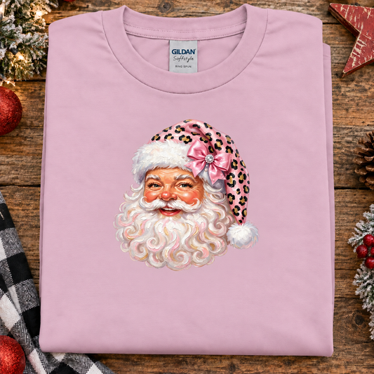 A folded light pink Gildan Softstyle t-shirt featuring a graphic of Santa Claus's face wearing a pink leopard-print hat with a bow, displayed on a rustic wooden background with holiday decorations.