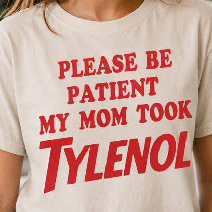 White t-shirt with red text 'PLEASE BE PATIENT MY MOM TOOK TYLENOL' worn by a person.