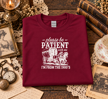 A folded cardinal red graphic T-shirt features a white printed graphic that reads 'please be PATIENT with me I'M FROM THE 1900's' with an illustration of a horse-drawn carriage.