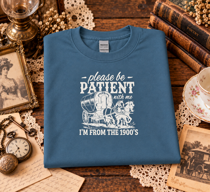 A folded indigo blue graphic T-shirt features a white printed graphic that reads 'please be PATIENT with me I'M FROM THE 1900's' with an illustration of a horse-drawn carriage.