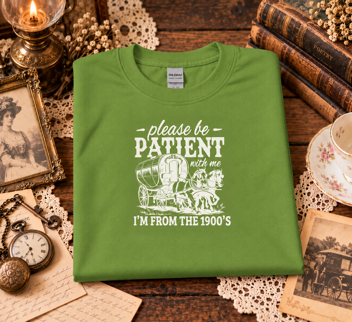 A folded kiwi green t-shirt with a white graphic print reading 'please be PATIENT with me I'M FROM THE 1900's' with a vintage covered wagon illustration, displayed on a rustic wooden table surrounded by antique-style props including books, a candle, a pocket watch, and a teacup.