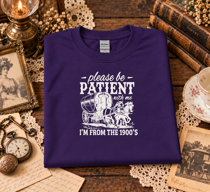 A folded purple t-shirt with a white graphic print reading 'please be PATIENT with me I'M FROM THE 1900's' with a covered wagon illustration, displayed on a rustic wooden table surrounded by vintage props like books, a candle, a pocket watch, and a teacup.