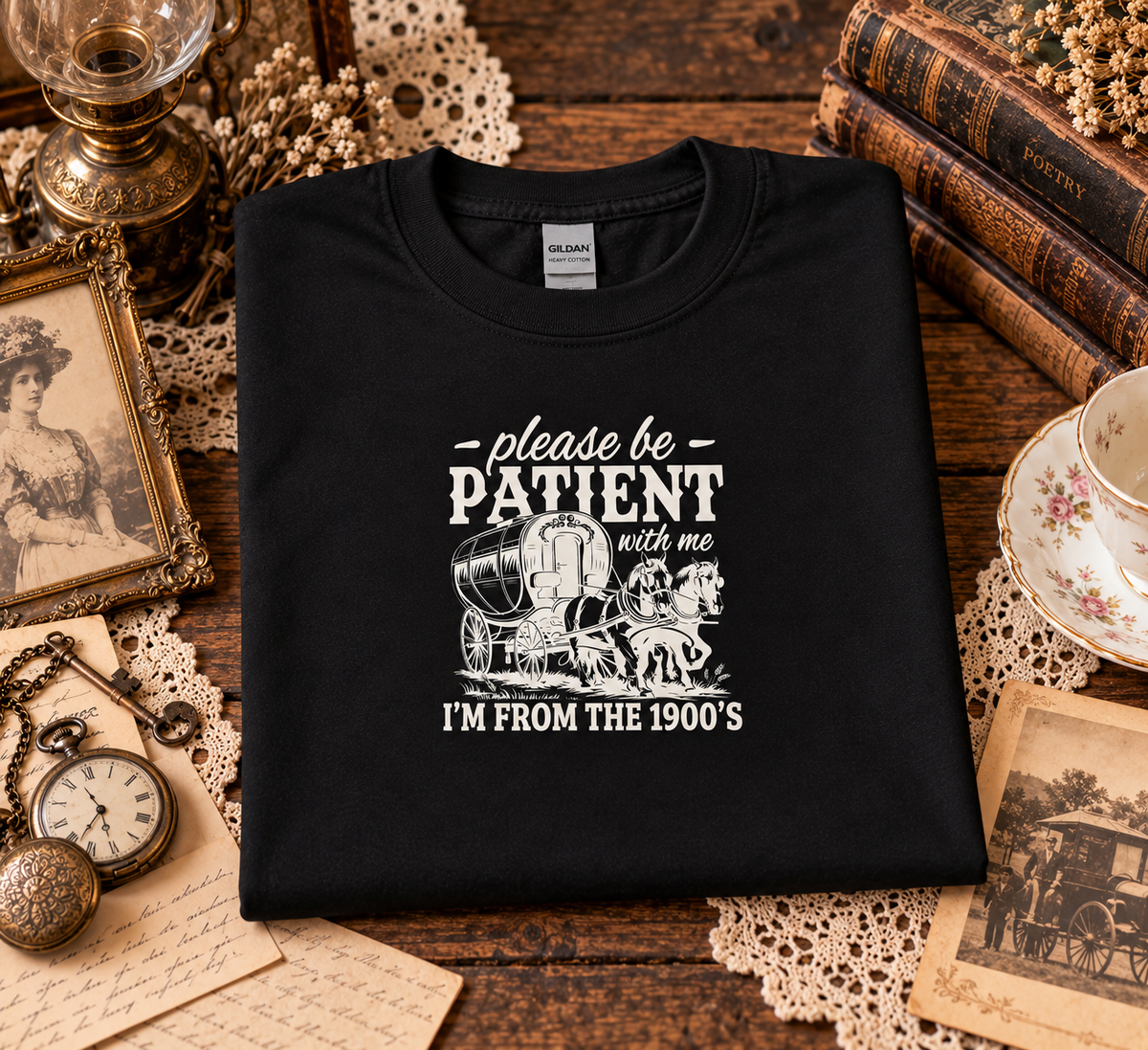 A folded black t-shirt with a white graphic print reading 'please be PATIENT with me I'M FROM THE 1900's' with an illustration of a covered wagon, displayed on a rustic wooden table surrounded by vintage props including books, a teacup, a pocket watch, and lace doilies.